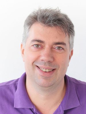 Porträt von Zahnarzt Dr. Suicu aus Flensburg in lila Polo-Shirt, lächelnd vor weißem Hintergrund.